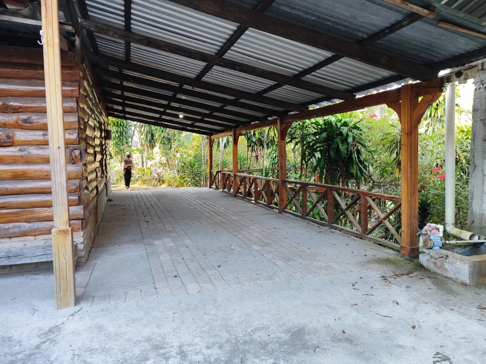 Área techada lateral de cabaña de madera con pasillo amplio y vista a jardín tropical